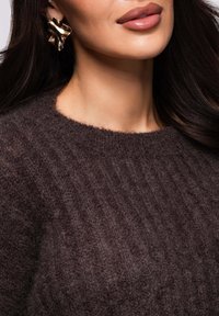 Mujer con suéter oscuro y peludo y un gran pendiente abstracto dorado, con labios brillantes de color malva y cabello largo y oscuro cayendo en cascada.