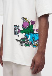 Personnage violet de dessin animé en sweat à capuche vert et casquette faisant du skateboard, tenant une caméra, avec un pantalon bleu et des baskets colorées sur un t-shirt blanc.