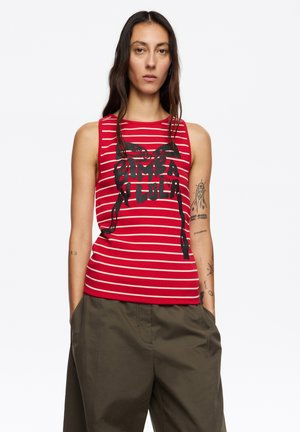 Top sin mangas con rayas rojas y blancas y texto gráfico en negro. Textura acanalada, escote redondo. Combinado con pantalones sueltos de color verde oliva.