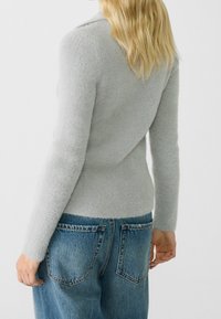 Persona bionda di spalle, vestita con un morbido maglione grigio chiaro e jeans di denim blu con tasche posteriori visibili, su sfondo bianco.