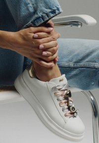 Zapatilla de cuero blanca con puntera redondeada, cordones de satén en rosa y adornos de piedras preciosas decorativas en los cordones. Suela texturizada, diseño minimalista.