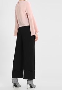 Vaaleanpunainen pusero, jossa on kellohihat, yhdistettynä leveälahkeisiin mustiin housuihin, joissa on valkoiset ompeleet. Mallilla on päällään ruudulliset korkokengät.