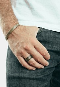 Bracciale in metallo con design texturizzato e intrecciato al polso, abbinato a un anello coordinato. Entrambi gli oggetti sembrano argentati, posizionati su fondo in denim.