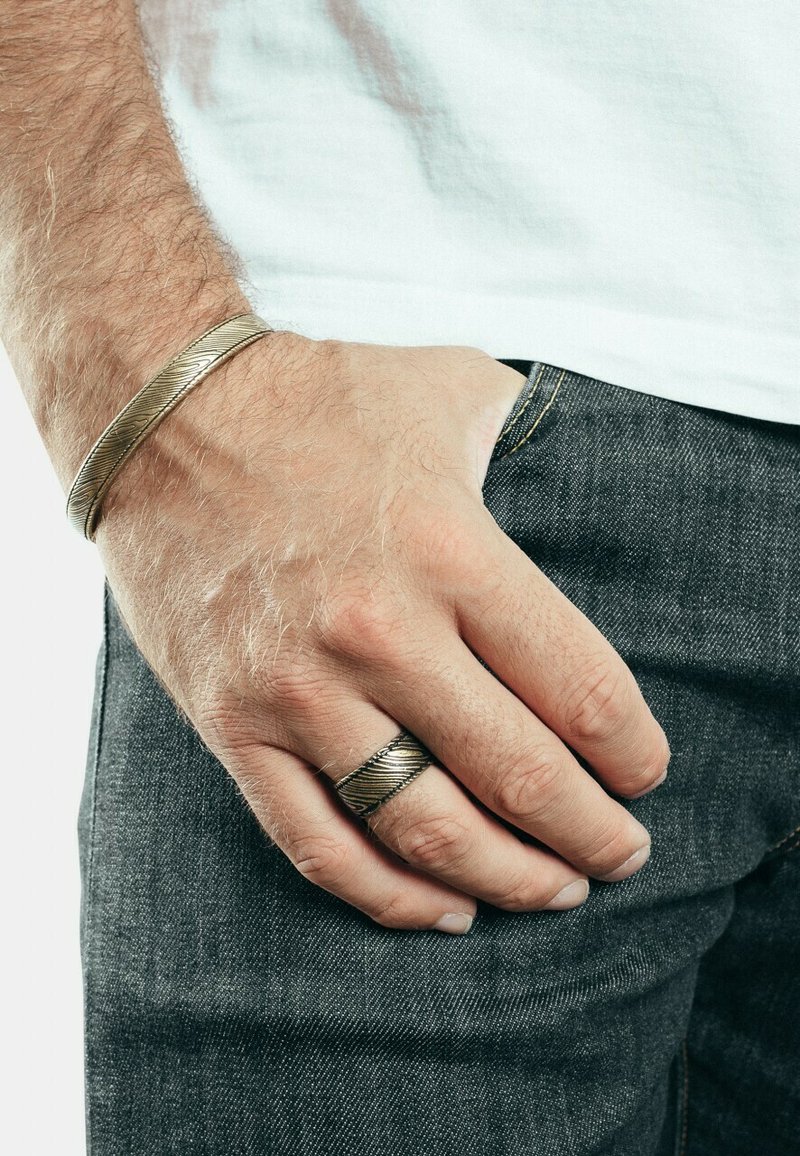 Bracciale in metallo con design texturizzato e intrecciato al polso, abbinato a un anello coordinato. Entrambi gli oggetti sembrano argentati, posizionati su fondo in denim.