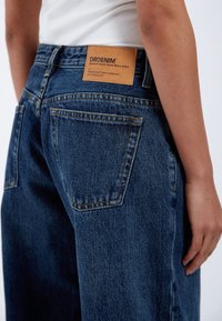 Mörkblå denimjeans med hög midja, med en brun läderetikett bak med märkesnamn, synlig sömnad och två bakfickor.
