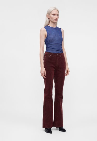 Femme grande aux cheveux blond clair portant un haut bleu sans manches, un pantalon bordeaux taille haute et des chaussures noires pointues, debout sur un fond blanc.