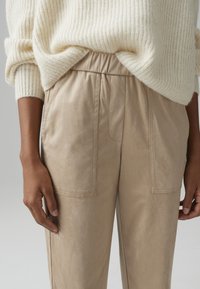 Beige faux leather pants with a relaxed fit, featuring an elastic waistband and two frontal pockets. Soft texture with subtle sheen.