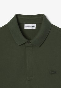 Polo shirt Lacoste verde scuro con colletto a costine, patta con tre bottoni e logo ricamato sul lato sinistro del petto. Vestibilità regolare, tessuto testurizzato.