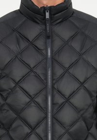 Giacca a sacco nera trapuntata con colletto alto e chiusura frontale con zip. Presenta motivi a diamante e una superficie liscia, leggermente lucida.
