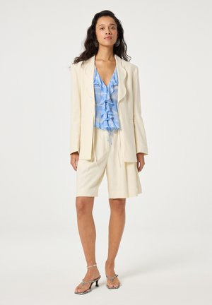 Vrouw draagt een crème blazer en shorts met een blauwe blouse met ruches, zilveren sandaaltjes met hakken en creooloorbellen, staand op een witte achtergrond.