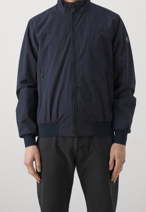 Man draagt een marineblauwe jas met rits, zakken met rits en elastische manchetten, gecombineerd met een zwarte broek tegen een effen achtergrond.