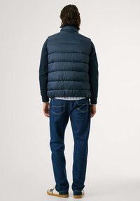 Marineblå puffervest med quiltet tekstur, brukt over en langermet skjorte og kombinert med mørke jeans og lyse sko. Bakfra.