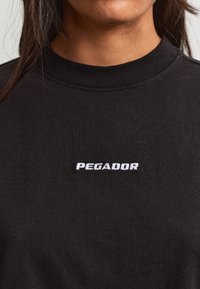Nahaufnahme einer Person, die ein schwarzes Rundhalsshirt trägt, auf dessen Brust das Wort "PEGADOR" in Weiß gestickt ist.
