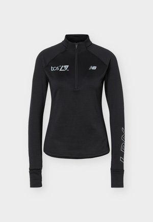 Μαύρο αθλητικό μπλουζάκι μακριάς μανίκας με φερμουάρ έως το ύψος του στήθους, λευκά λογότυπα "tcs L77 2026" και New Balance στο στήθος, ανακλαστικό "NB" στο αριστερό μανίκι.