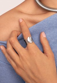 Silberner und schwarzer Metallring mit einer glatten, polierten Oberfläche in modernem Design. Wird an einem Finger mit einem hellen, gestrickten Oberteil getragen.