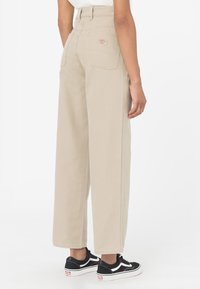 Béžové široké kalhoty s vysokým pasem, zadními kapsami a viditelným švem, doplněné černými teniskami s bílými akcenty.