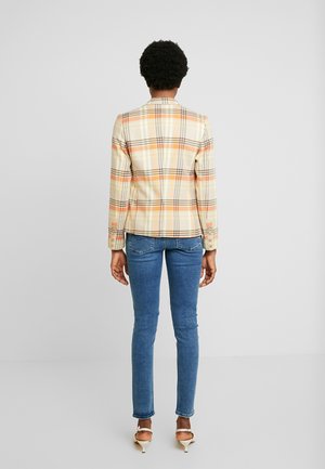 Osoba o krótkich kręconych włosach, ubrana w beżowo-pomarańczową kratkowaną marynarkę, niebieskie obcisłe dżinsy i beżowe szpilki, stoi tyłem.