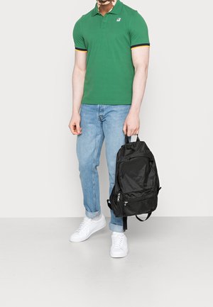 Homme portant un polo vert, un jean bleu clair, des baskets blanches, tenant un sac à dos noir par sa poignée supérieure, debout sur un fond uni.