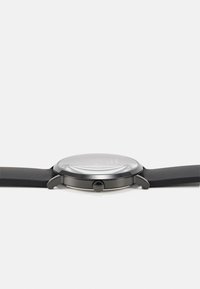 Round watch with a smooth, glass face and matte black silicone strap. Features a metallic case and a round crown on the side.