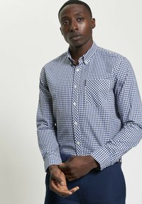 Camicia con bottoni in tessuto a quadretti blu e bianchi, maniche lunghe, taschino sul petto e colletto dritto. Il tessuto sembra liscio e leggero.