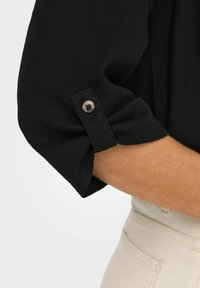 Schwarzes Strickhemd mit aufgerollten Ärmeln und Knopfdetail. Weicher Stoff und lässiges Design. Teil eines hellen Outfits.