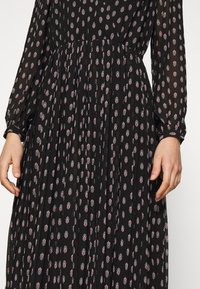 Zwart geplisseerd jurk met lange mouwen en een klein rood en wit bloemmotief, getoond van taille tot polsen met ontspannen handen aan de zijkanten.