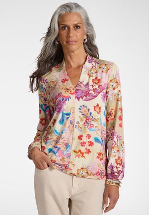 Mujer madura con cabello largo y gris, vistiendo una blusa floral colorida y pantalones beige, con una mano en el bolsillo, fondo neutro.