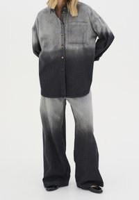 Camisa de mezclilla y pantalones acampanados en un degradado de negro a gris. La camisa tiene un bolsillo en el pecho y botones; los pantalones son de corte suelto.