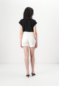 Top negro de manga corta combinado con pantalones cortos blancos de cintura alta, con una textura suave y dos bolsillos traseros. La modelo está de espaldas.