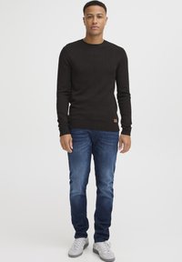 Zwarte gebreide trui met lange mouwen, ronde halslijn, gecombineerd met blauwe denim jeans en grijze sneakers, met een klein bruin leren accent.