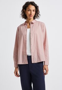 Donna che indossa una camicia a righe rosa e bianca sopra una maglietta bianca, abbinata a pantaloni blu navy scuri, in piedi su uno sfondo bianco.