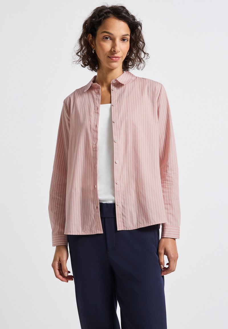 Donna che indossa una camicia a righe rosa e bianca sopra una maglietta bianca, abbinata a pantaloni blu navy scuri, in piedi su uno sfondo bianco.