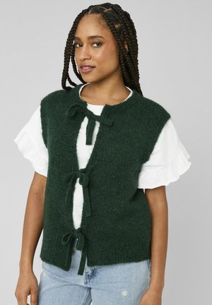 Femme aux cheveux tressés portant un gilet en maille vert foncé noué par trois nœuds par-dessus un haut blanc à manches courtes à volants et un jean bleu clair.