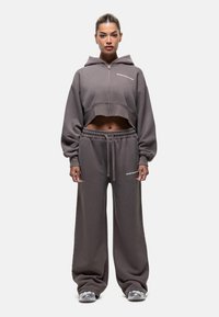 Bruine crop hoodie met rits en logo op de borst, gecombineerd met losse joggingsbroeken met een elastische tailleband. De sweatpants hebben zijzakken.