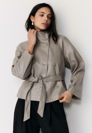 Frau, die eine taupefarbene Lederjacke mit hohem Kragen, Knopfverschlüssen und gebundenem Gürtel anpasst, schwarze Hose und silberne Creolen trägt.