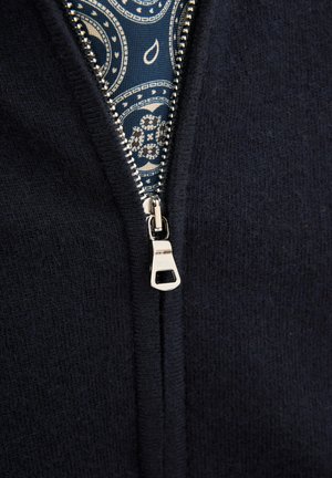 Marineblaue Jacke mit Reißverschluss aus einem weichen Material, ausgestattet mit silbernen Reißverschlussbeschlägen und einem gemusterten blauen Futter, das am Öffnungsbereich sichtbar ist.