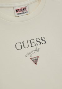 Camiseta de algodón color crema con el logotipo bordado "GUESS" en negro y "originals" en cursiva, con un acento de triángulo rojo.