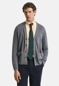 Grauer Cardigan mit V-Ausschnitt, fünf Knöpfen, seitlichen Taschen und gerippten Bündchen. Getragen über einem gelben Hemd und einer grünen Krawatte mit kleinen goldenen Punkten.