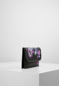 Borsa a tracolla in pelle nera con ricami floreali colorati, bordi a punto catena e chiusura a patta, esposta su un piedistallo bianco.