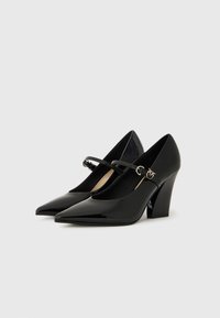 Escarpins en cuir verni noir à talon aigu, avec un bout pointu et une sangle sur le coup de pied, ornés de détails en métal.