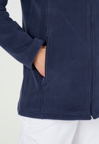 Chaqueta de forro polar azul oscuro con un bolsillo lateral con cremallera, textura suave y cuello alzado. Está diseñada para ofrecer comodidad y calidez.