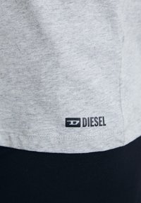 T-shirt en coton gris avec un logo "DIESEL" noir sur l'ourlet. Le tissu a une texture douce avec des variations subtiles de ton.
