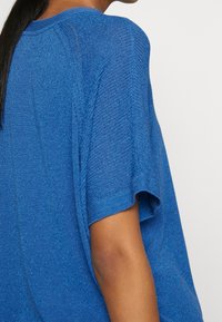 Modrý pletený top volného střihu, s krátkými rukávy a texturovaným povrchem. Tkanina má jemný třpytivý efekt, který zvyšuje její vizuální přitažlivost.