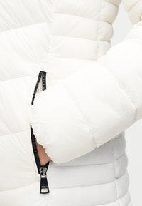 Λευκό μπουφάν με φουσκωτό ύφασμα, πλευρική τσέπη με φερμουάρ, κατασκευή με ραφές και απαλή υφή. Τα μανίκια είναι ελαφρώς φουσκωτά για άνεση.