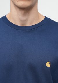 Navy sininen puuvillapaita pyöreällä kaula-aukolla ja ribbausreunalla. Yhdessä vasemmassa rinnassa on pieni kultainen brodeerattu logo.