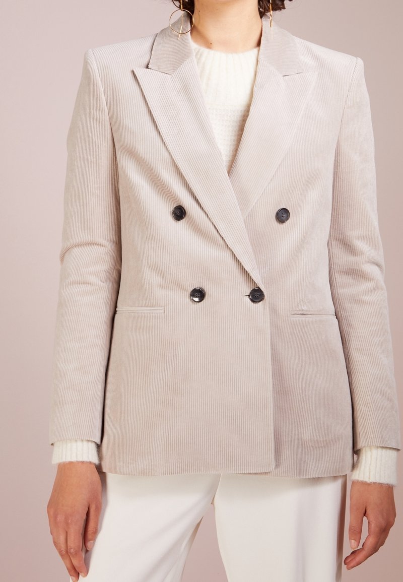 Hellbeige Cordjacke aus Rippstoff mit doppelter Knopfleiste, Reverskragen, zwei schwarzen Knöpfen und seitlichen Taschen, kombiniert mit einem cremefarbenen Pullover.