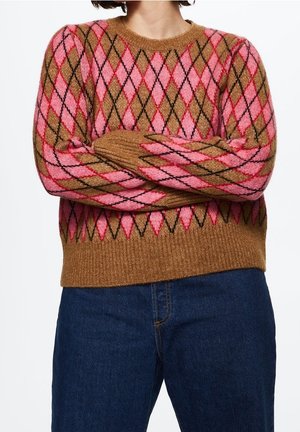 Personne portant un pull marron et rose à motif losanges, les bras croisés, associé à un jean en denim bleu foncé.