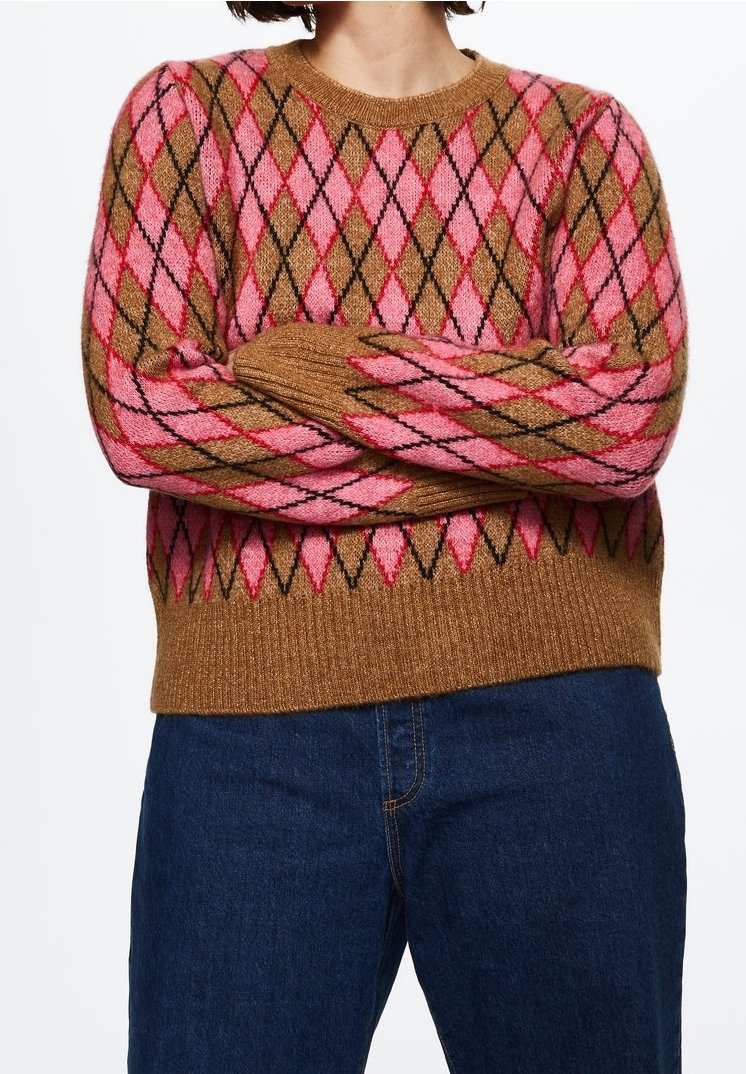 Personne portant un pull marron et rose à motif losanges, les bras croisés, associé à un jean en denim bleu foncé.