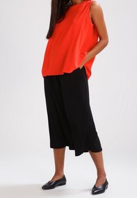 Červený top bez rukávů s asymetrickým lemem v kombinaci s černými širokými kalhotami. Model má na sobě černé ploché boty; minimalistický design, hladká textura.