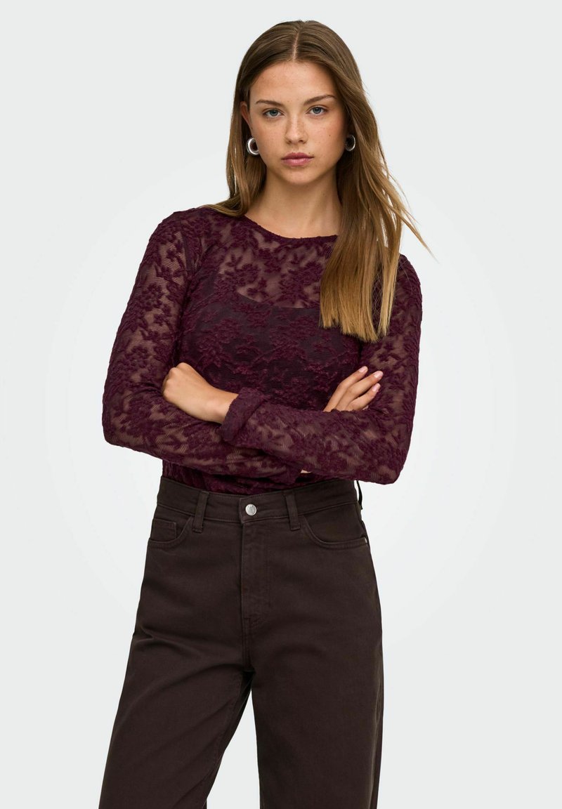 Jeune femme aux longs cheveux châtain clair porte un top en dentelle bordeaux transparent et un pantalon sombre taille haute, debout, les bras croisés devant un fond uni.